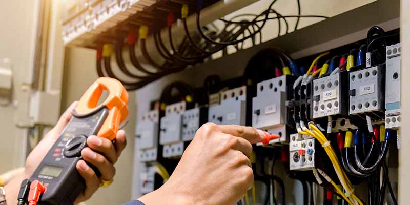 Foto do interior de um painel elétrico com diversos componentes e uma mão segurando um multímetro.